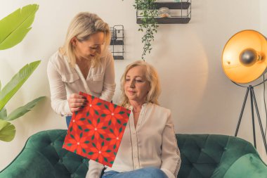 Mutlu olgun anneyi kutlayan ve doğum günü hediyesi veren gülümseyen büyük anne, Anneler Günü 'nde yetişkin torunundan sürpriz aldığı için heyecanlı.