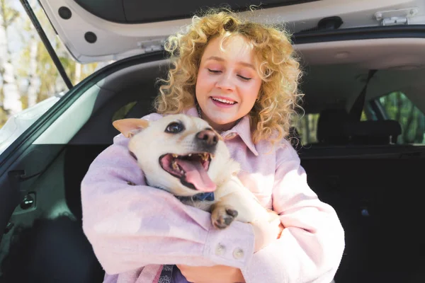 Happy female dog owner in a pink jacket with her animal companion sitting in the back of car, having fun. Outdoor shot. — Zdjęcie stockowe