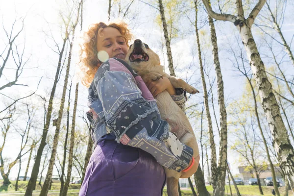 Utomhus fotograferad av en glad ung kvinna med kinky hår som står nära träd och håller en glad hund i sina armar. — Stockfoto