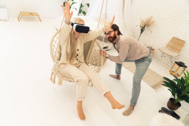 Minimalist bir apartman dairesinde sıradan kıyafetler giyen ve sanal gerçeklik gözlükleriyle oynayan genç bir çift..