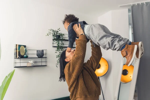 Little multiracial toddler boy lifted by his strong young mum in their modern apartment. Happy childhood concept. — Stock fotografie