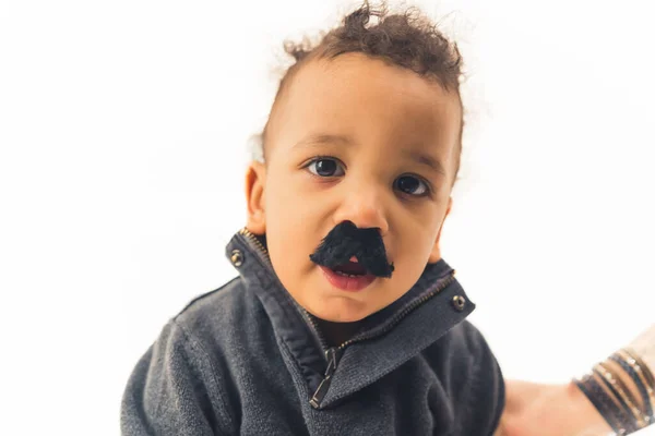 Portrait studio d'un adorable bébé garçon biracial aux cheveux bouclés foncés regardant la caméra tout en portant une fausse moustache noire sur fond blanc. — Photo