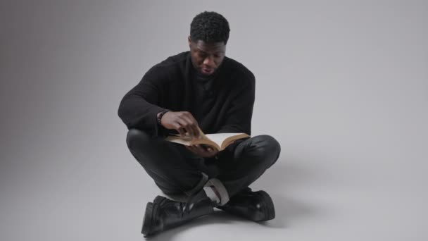 Focused interested black guy wearing black clothes sitting cross-legged on the floor reading a book, turning pages, looking up. — Vídeo de Stock