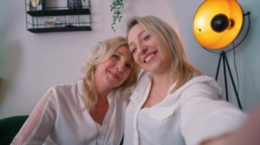 Indoor shot of two related caucasian women in their 40s wearing white elegant shirts takinga selfie, hugging each other and smiling.