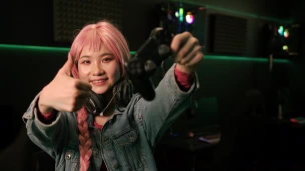 Young asian cute happy female player with pink hair and a long braid wearing headphones around her neck, holding a gamepad, smiling, showing thumbs up sign. — Stockvideo