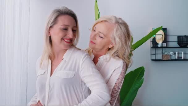 Jedna žena s vlnitými blond vlasy, stojící vzadu a objímající jinou bělošku středního věku. Oba se smějí a baví v elegantním bytě. — Stock video
