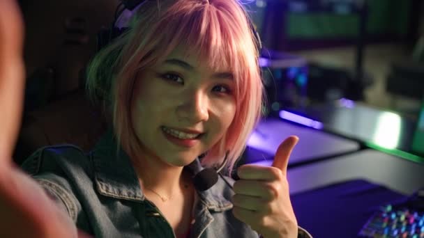 Close-up indoor shot of a beautiful asian pink-haired gamer girl sitting in a swivel gaming chair in a room with many colorful keyboards and computers doing a selfie. — 비디오