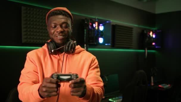 Cheerful african american male gamer dressed in an orange outfit, with big black headphones around his neck, holding a gamepad in his hands, smiling. — Stok video