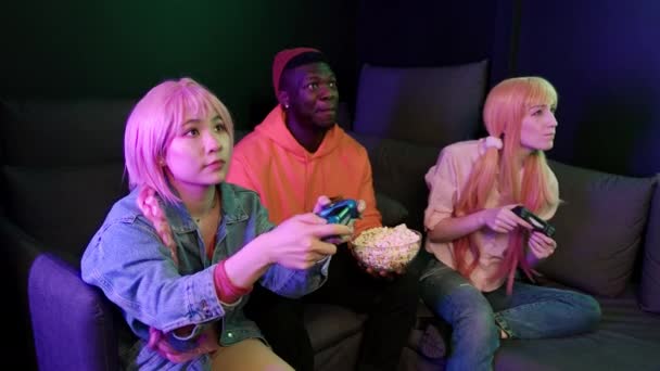 Fotografía interior de un grupo de milenarios interraciales jugando videojuegos, sentados en un sofá oscuro. Asiática adolescente winnig una competencia y animando. — Vídeo de stock