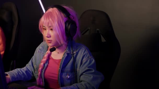 专注的亚洲女性游戏玩家，戴着粉色长辫子，身穿斜纹棉布夹克，凝视着屏幕，在视频游戏的在线流媒体中聚精会神地观看. — 图库视频影像