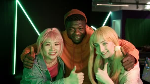 Multiracial young cheerful friends standing close together posing at camera in a dark gaming room with neon lights showing peace and thumbs up signs. — Stockvideo