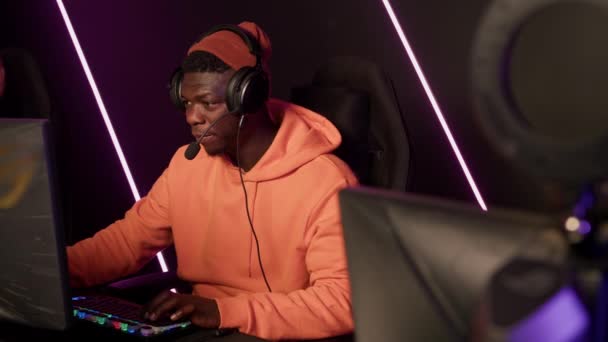 Focused african american male player wearing a beanie and a sweatshirt, sitting in a comfortable gaming armchair in front of a giant computer screen. — Stock videók