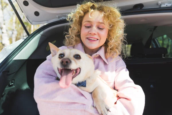 Charming blonde European girl hugging her cute dog while sitting in the car trunk medium shot outdoor pets and humans concept — Stockfoto
