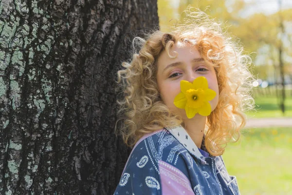 Blonde curly-haired young Caucasian girl holding a flower in her mouth in the park medium closeup outdoors — Zdjęcie stockowe