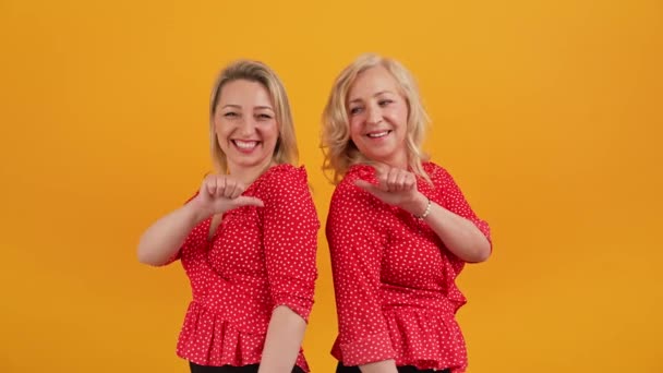 Deux jolies jolies femmes d'âge moyen se pointant dos à dos avec leurs pouces. Studio tourné sur fond orange. — Video