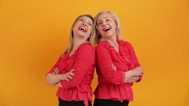 Mittlere Studioaufnahme auf gelbem oder orangefarbenem Hintergrund mit zwei erwachsenen kaukasischen Freundinnen, die Rücken an Rücken stehen, die Arme verschränkt zueinander halten und laut lachen. — Stockvideo