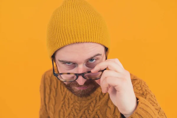 European unshaven confused suspicious guy slipping off his eyeglasses off the nose raising his eyebrow looking directly at the camera. Studio shot, yellow background. — ストック写真