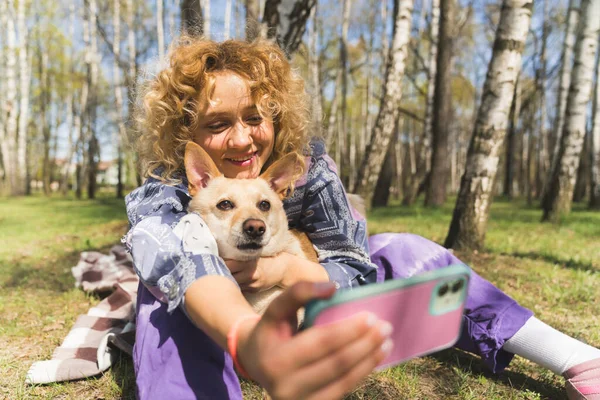 Close-up shot of a joyful beautiful caucasian curly-haired girl sitting on the grass in a park holding a mobile phone, taking a picture of her and her dog. — Zdjęcie stockowe