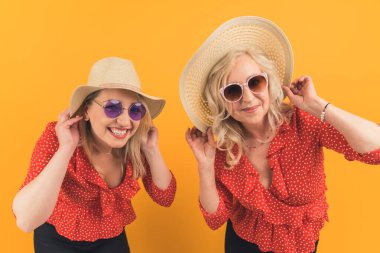 two attractive women together having fun, mother and daughter in similar clothes and hats smiling and rejoicing over orange background