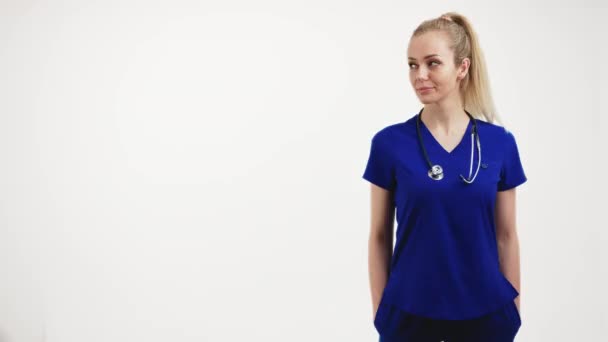 Studio shot over white background of a young pale blonde attractive doctor in blue scrubs with a stethoscope. Medical concept — Vídeo de stock