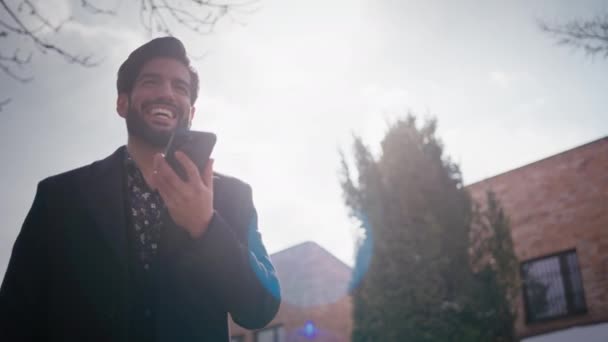 Excited cheerful latin guy in his 30s standing and speaking on a speaker on a mobile phone on a sunny day. Outdoor medium shot — Wideo stockowe