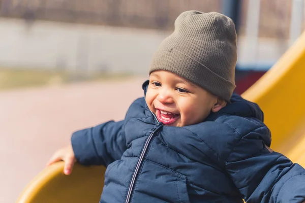 Outdoor portrait shot of a young biracial baby boy in a hat and warm coat smiling and sliding on the playground slide. — 图库照片