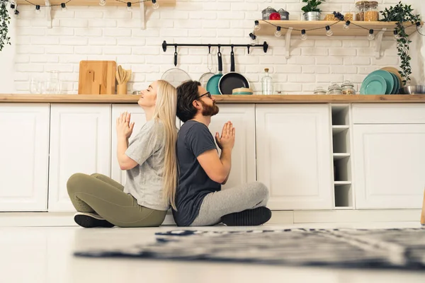 Jovem casal adulto sentado no chão de volta para trás meditando juntos em uma cozinha acolhedora bem equipada. Tiro interior — Fotografia de Stock