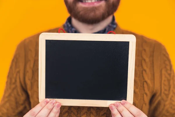 Cheerful bearded millennial guy in a casual outfit holding a tiny blank black board. Studio shot on orange background, focus on the foreground. — Φωτογραφία Αρχείου