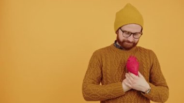 young bearded man with glasses holding a fake heart in front of his chest medium closeup orange background studio shot copy space 