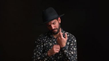 Siyah arkaplanda, siyah şapkalı ve çiçekli gömlek giyen yakışıklı Latin sakallı bir adamın gömlek manşetini düzeltirken orta boy bir fotoğraf.. 