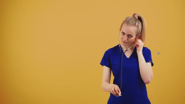 Bionda concentrata medico femminile nei suoi 20 anni indossando camici blu tenendo il suo stetoscopio e l'ascolto. Studio medio girato su sfondo arancione — Video Stock