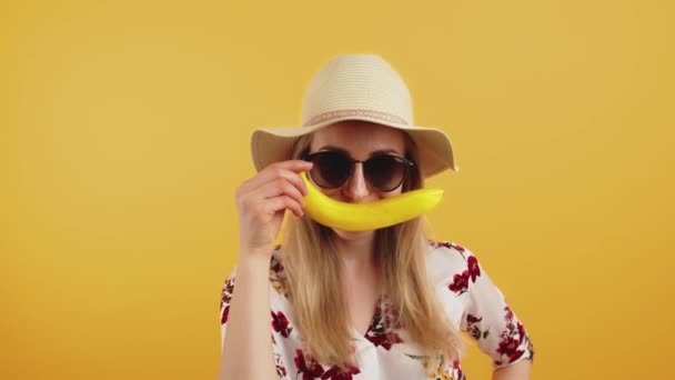 20 대 유럽 여성, 여름 모자, 큰 선글라스, 꽃이 달린우아 한 셔츠를 입고 입에 미소를 머금은 채긴 노란 바나나를 들고 있다.. — 비디오
