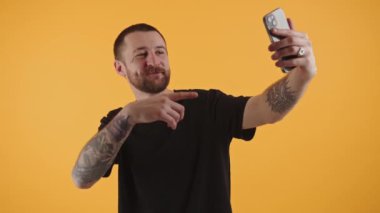 Yakışıklı sakallı adam akıllı telefonuyla video çekiyor. Kolunu hareket ettiriyor. Stüdyo sarı arka plan. 