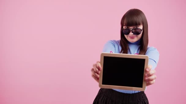 Mignon hipster européen brunette adolescent dans lunettes de soleil montrant maquette tableau noir pour caméra sur fond rose. — Video