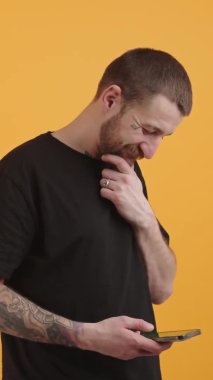 Dikey video, dövmeli bir adamın telefonuna yapışmış, kameraya bakıyor ve sarı arka plandaki izole stüdyo çekimine gülüyor. 