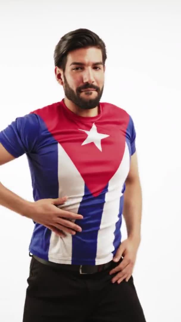 Un joven barbudo seductor y guapo con una camiseta con una bandera cubana, bailando, moviendo las manos. Estudio vertical sobre fondo blanco. — Vídeo de stock