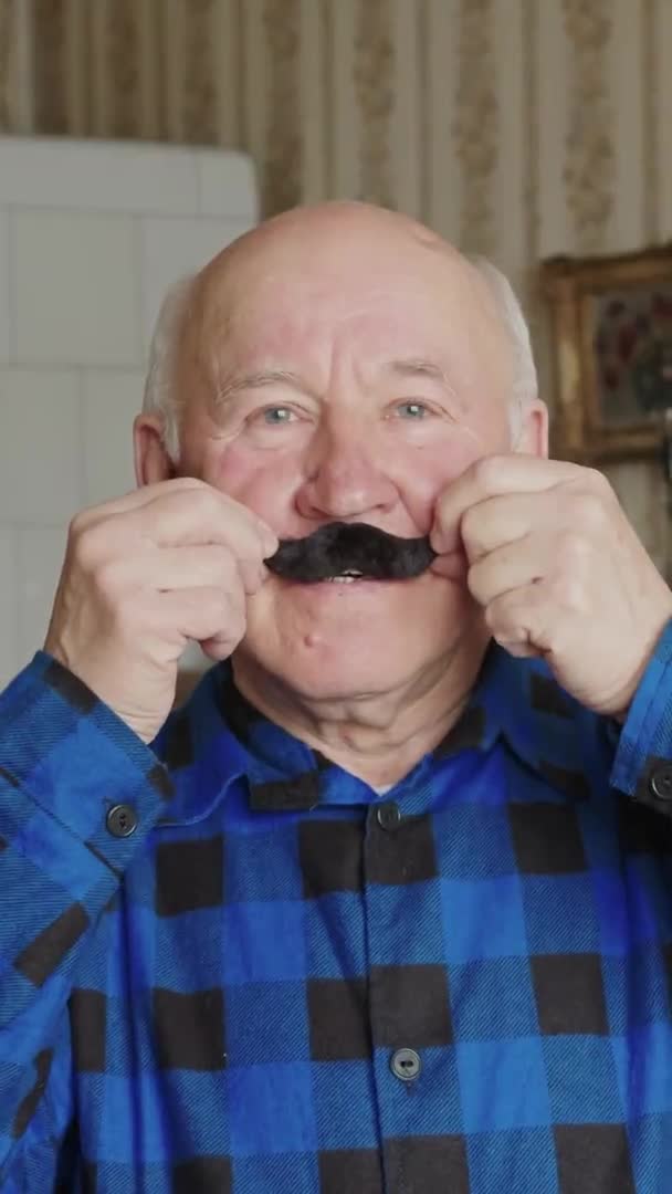 Χαρούμενος ηλικιωμένος Ευρωπαίος προσαρμόζει ψεύτικο μουστάκι κάθετο βίντεο μεσαίο closeup έννοια εσωτερικό ηλικιωμένους — Αρχείο Βίντεο