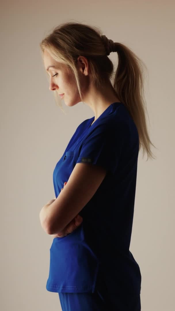 Вертикальне відео красивої блондинки медсестри в синьому костюмі середній знімок студії знімок світлого фону — стокове відео