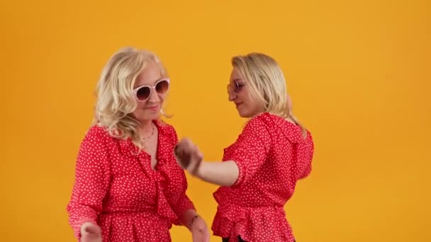 Två europeiska blonda tusenåriga kvinnor i röda prickiga skjortor och solglasögon dansar, sjunger och har roligt. Studio skott på orange bakgrund. — Stockvideo