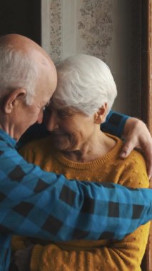 Mutlu yaşlı bir çift pencerenin önünde birbirlerine sarılıyor. Dikey video, kapalı mekan aşkı ve yaşlılar konsepti destekliyor.