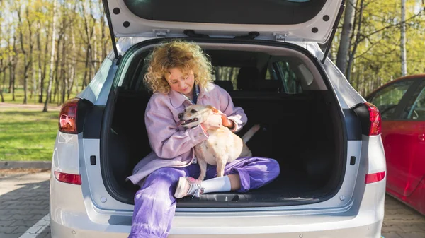 Nastolatka kaukaska dziewczyna z kręconymi włosami pieszczoty jej ładny pies podczas siedzenia w samochodzie, piękne zdjęcie w parku — Zdjęcie stockowe