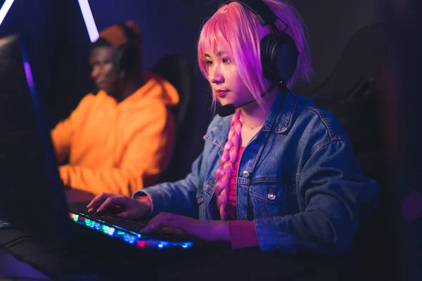 Молодая азиатка в наушниках и розовом парике играет в видеоигры, афроамериканец на заднем плане — стоковое фото