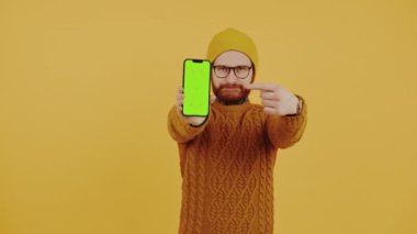 Sarı bereli, turuncu kazaklı, beyaz sakallı komik hippi adam yeşil ekran telefonunu işaret ediyor. Sarı arka plan stüdyo çekimi.