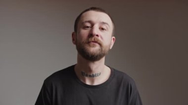 Siyah tişörtlü, sakallı beyaz bir adam kameraya bakıyor ve kızgın küstah yüz ifadesiyle başını sallıyor..