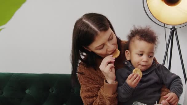 Αφρο-αμερικανική σγουρά μαλλιά μικρό παιδί και η μαμά του τρώνε μπισκότα στο πράσινο καναπέ σαλόνι φόντο αντίγραφο χώρο — Αρχείο Βίντεο