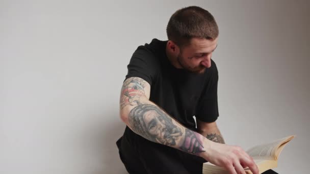 Fiatal szakállas kaukázusi tetovált férfi srác olvasás és essek oldalak a kedvenc könyve szürke háttér. — Stock videók