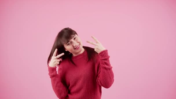 Crazy happy kaukasische junge erwachsene brünette Mädchen in rotem Pullover zeigt V-Zeichen in die Kamera und wedelt mit ihren Haaren herum. Isoliertes Studio auf rosa Hintergrund. — Stockvideo