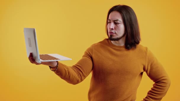 Estudio de toma de hombre caucásico confuso serio de mediana edad con una barba y pelo largo marrón de pie en el estudio en suéter amarillo cálido con su computadora portátil abierta en una mano sobre el fondo amarillo. — Vídeos de Stock