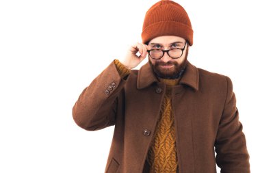 Bereli genç beyaz erkek komedyen ve sonbahar kıyafetleri gözlüklerine dokunuyor ve beyaz arka planda kameraya anlamlı bir şekilde bakıyor..