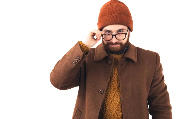 Jeune comédien caucasien masculin en bonnet et vêtements d'automne touchant ses lunettes et regardant suggestivement la caméra sur fond blanc. — Photo